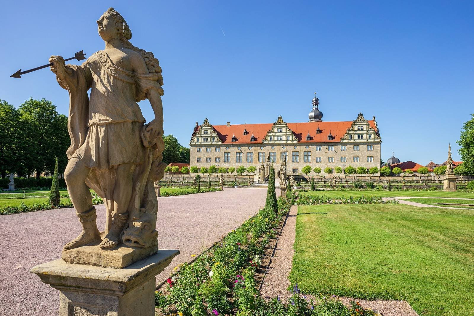 A Baroque statue in the foreground of the Weikersheim palace garden, with lawns, flower beds and the palace beyond